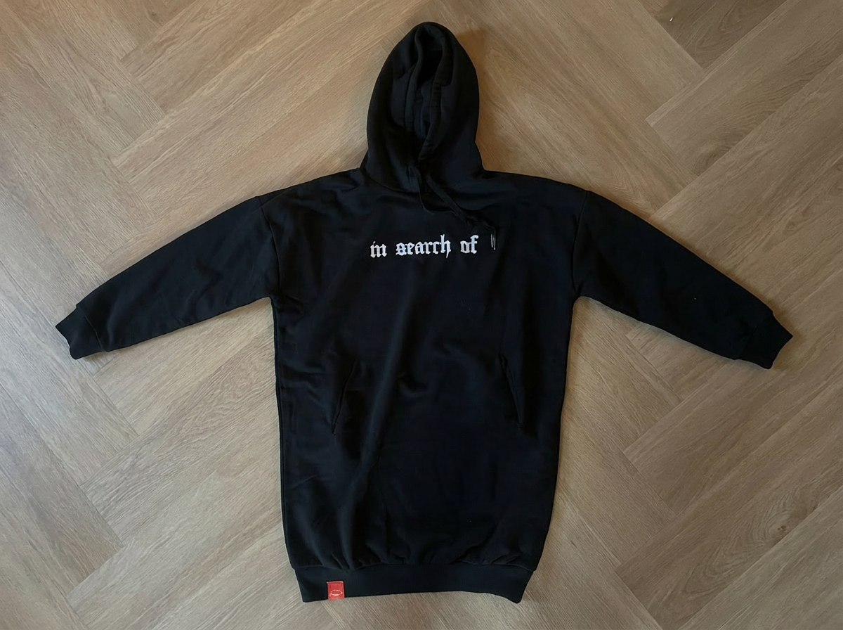 In Search Of Hoodie Dress black oversized – gothic "in search of" lettering on chest, red woven label at hem, flatlay on wood floor